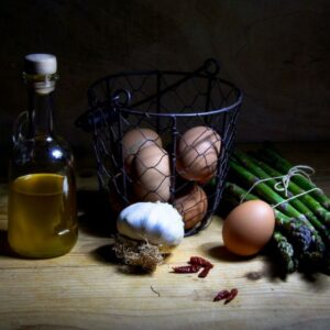Bottle of golden oil beside eggs, garlic, and asparagus on a wooden table in soft, moody light.