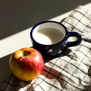 Apple and a navy mug in soft morning light on a checked cloth—a simple cue for whole food and hydration.