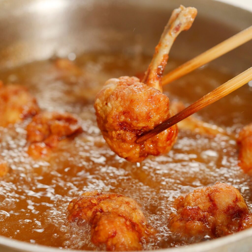 Chopsticks lifting a browned chicken bite from sizzling tallow in a pan