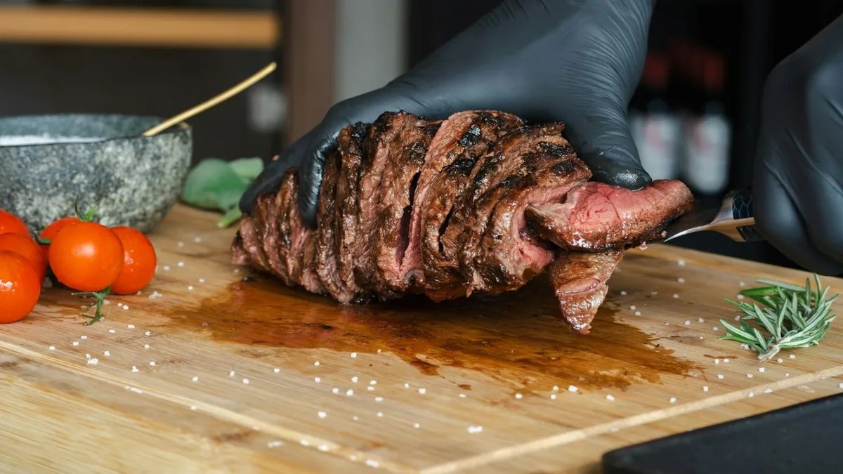 Sliced ribeye on a wooden board with gloved hands and juices.
