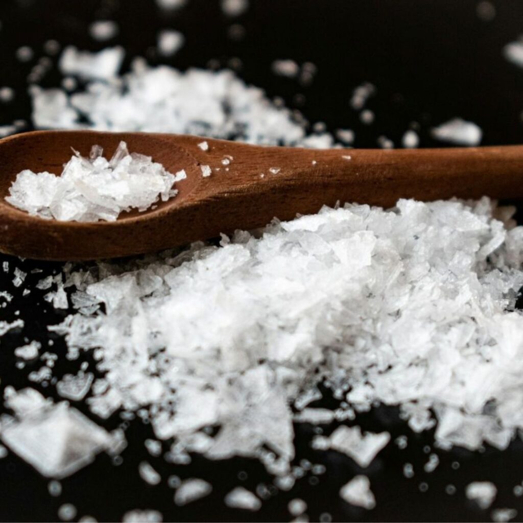 Sea salt flakes in a small wooden spoon on a dark surface.
