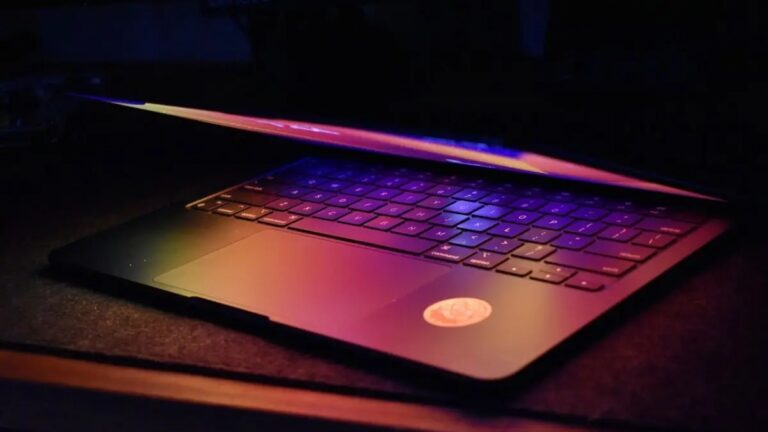 Dim evening laptop partly closed, purple-blue screen glow in a dark room.