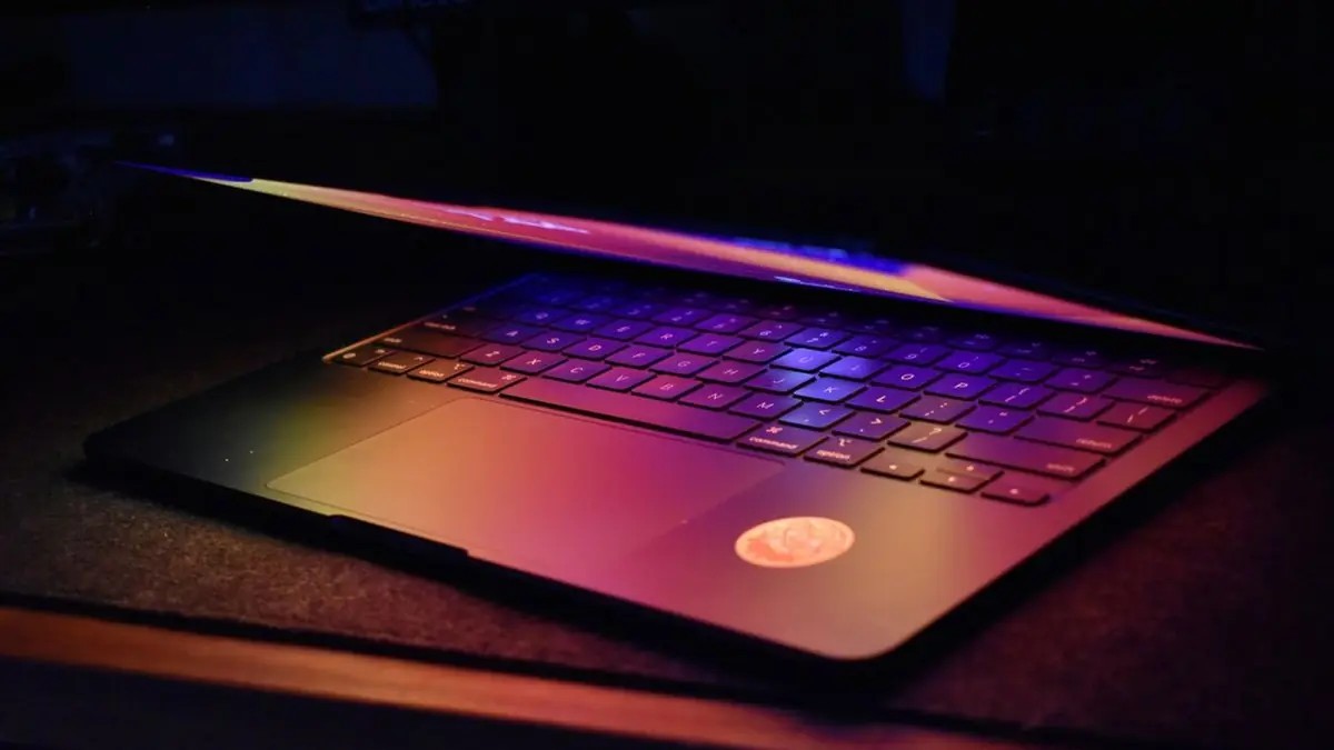 Dim evening laptop partly closed, purple-blue screen glow in a dark room.