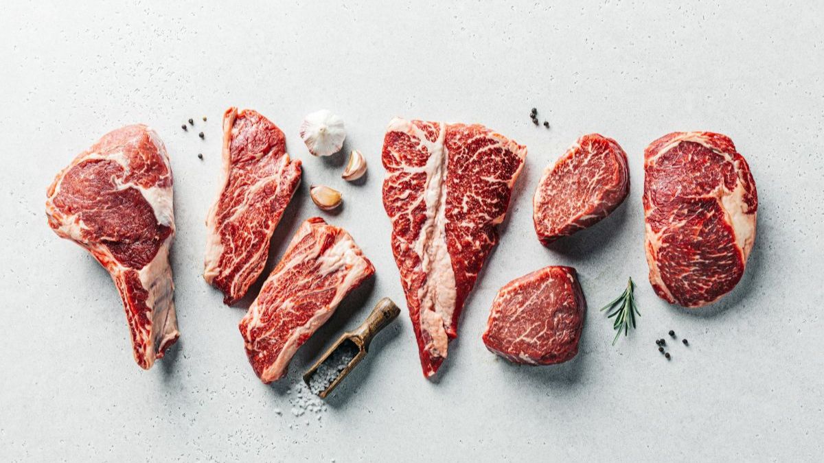 Assorted raw marbled beef cuts arranged in a row on a light surface with garlic, peppercorns, rosemary, and coarse salt.