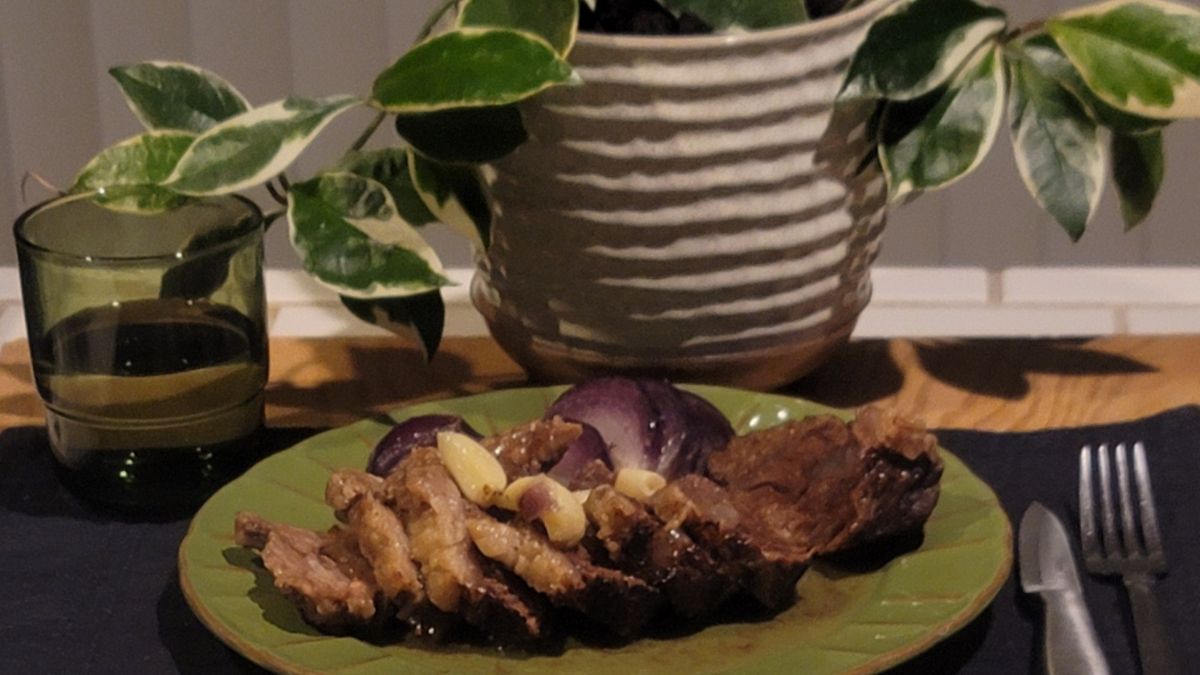 Sliced slow-cooked beef cheek fillet with garlic cloves and red onion on a green plate, with a green glass and leafy potted plant in the background.