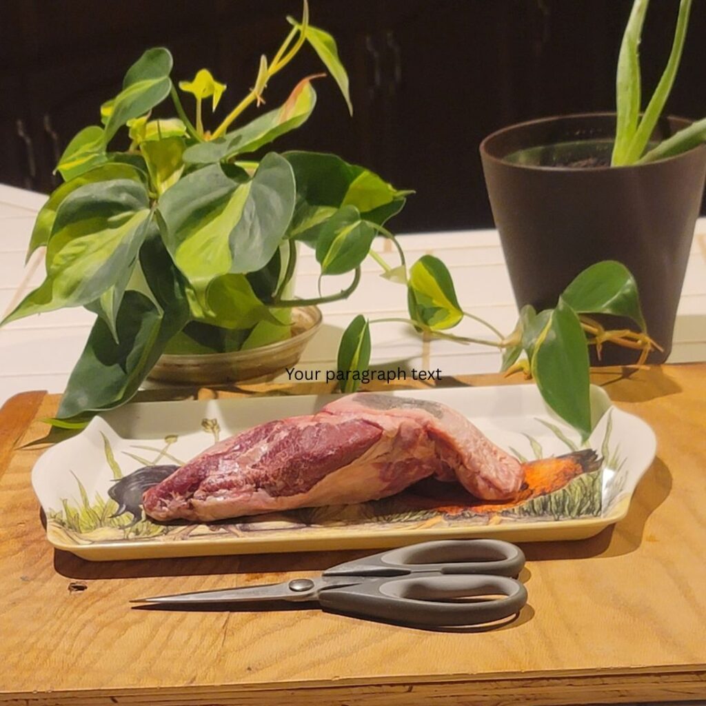 Raw grass-fed beef cheek fillet on a decorative tray with kitchen shears, surrounded by green houseplants on a wooden board.