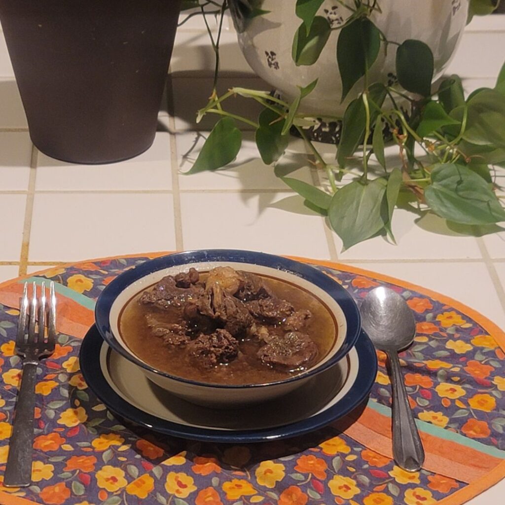 Bowl of chuck meat soup with rich broth on a floral placemat, set on a tiled countertop with a fork, spoon, and trailing houseplant nearby.