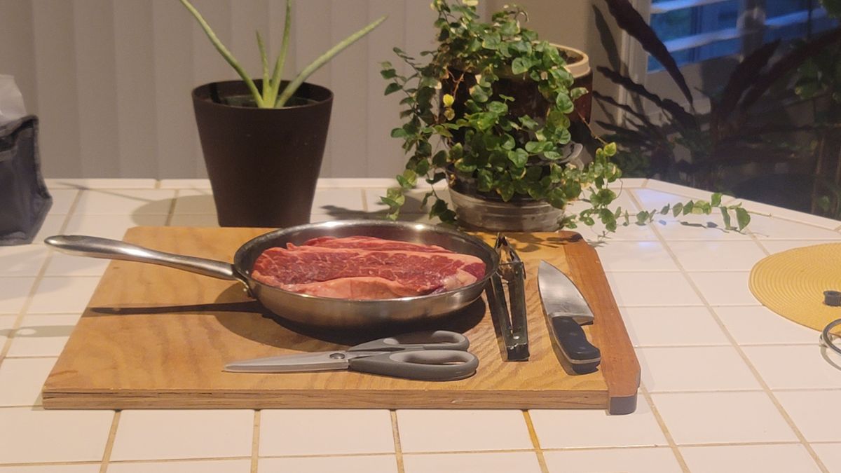 Raw chuck steaks in a stainless-steel pan on a wooden board with scissors, tongs, and a knife, surrounded by potted plants on a tiled countertop.