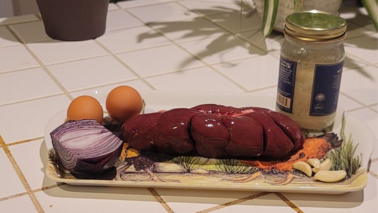 Grass-fed beef kidney, eggs, red onion, garlic and beef tallow arranged on a tray on a tiled kitchen counter.