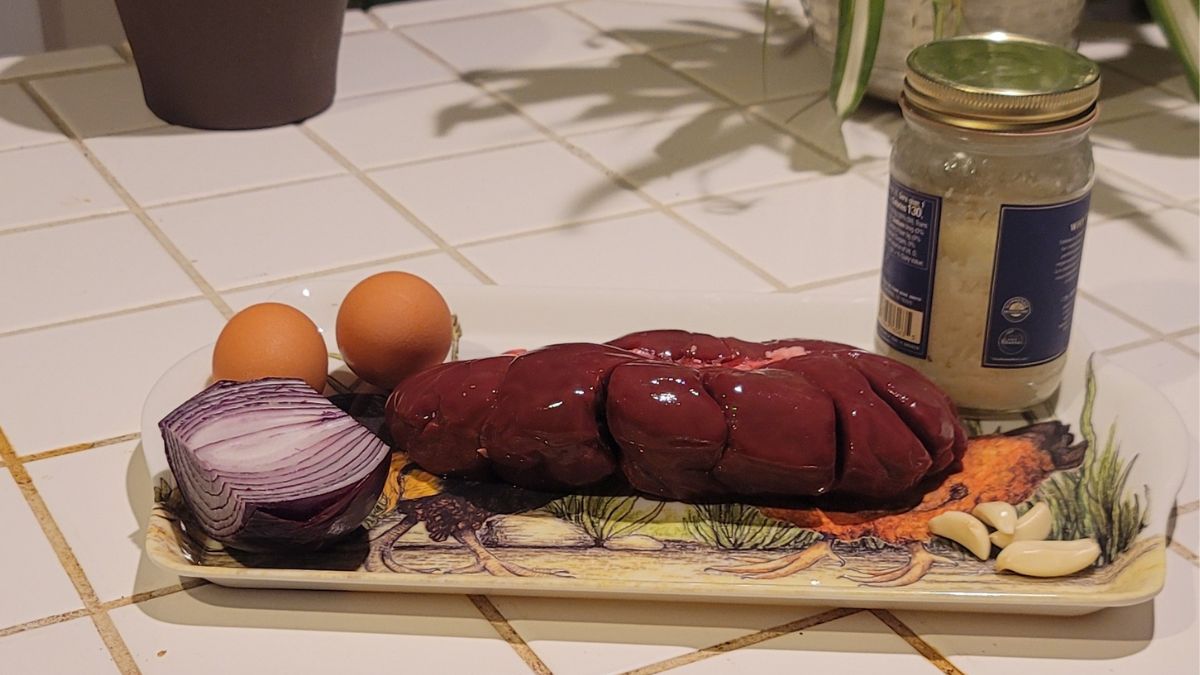 Grass-fed beef kidney, eggs, red onion, garlic and beef tallow arranged on a tray on a tiled kitchen counter.
