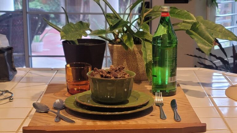 Bowl of oxtail and beef heart soup on a wooden board with sparkling water, cutlery, and green plants in the background