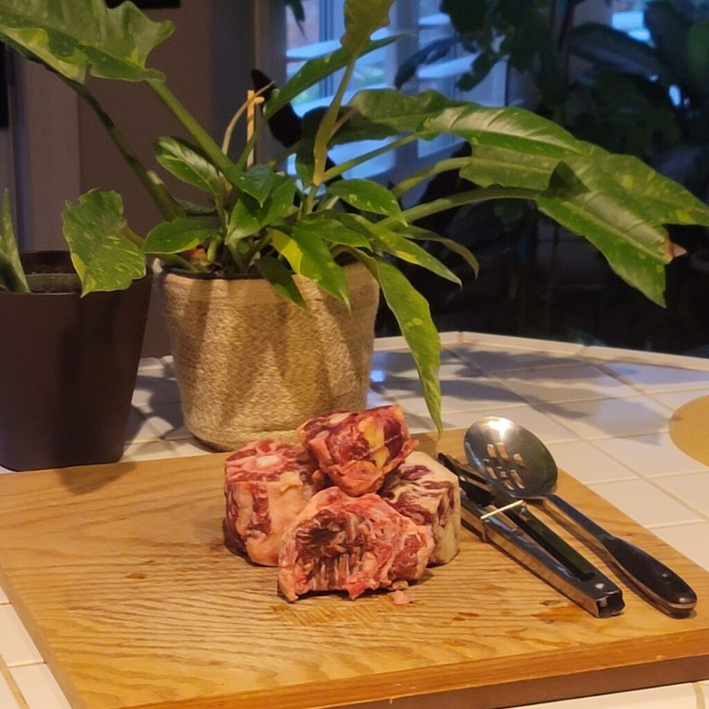 Raw oxtail pieces stacked on a wooden board with serving utensils and green houseplants behind