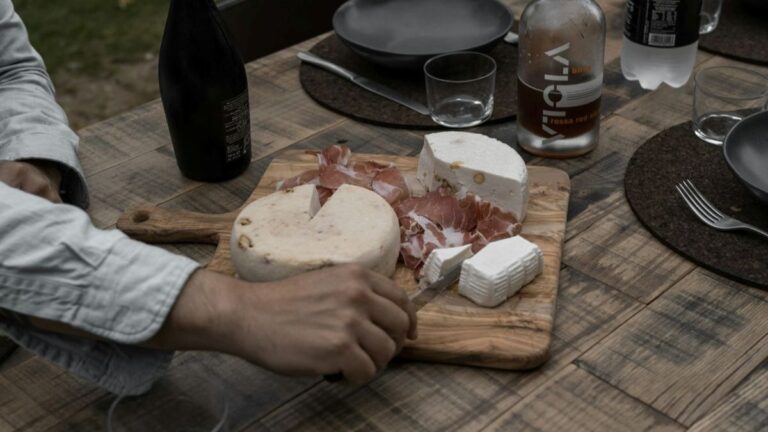 hand reaching toward a wooden board with cheese and cured meat on a rustic dinner table