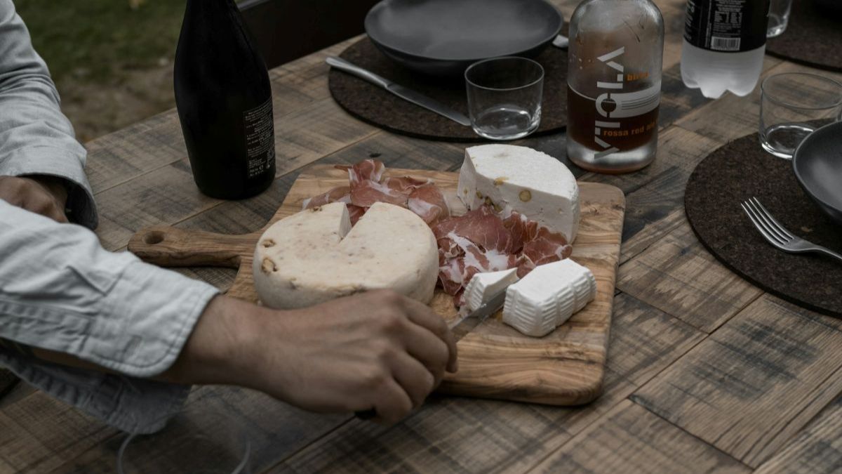 hand reaching toward a wooden board with cheese and cured meat on a rustic dinner table