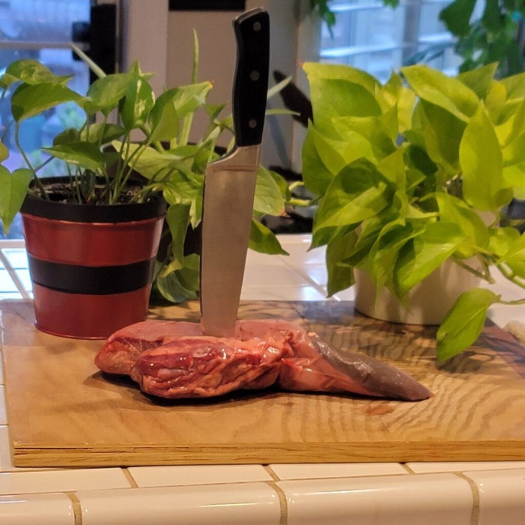 Raw beef tongue on a wooden board surrounded by kitchen plants.