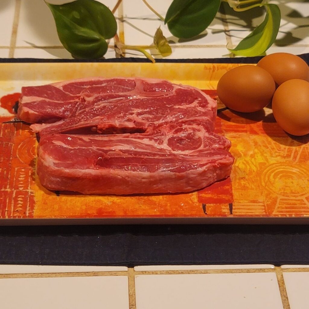 Raw lamb shoulder chops and whole eggs on an orange/red tray before cooking.