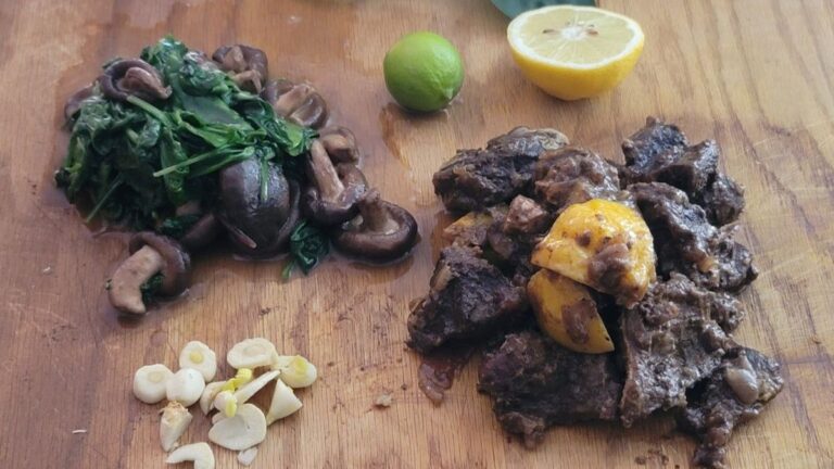 Cooked cheek meat with leafy greens, mushrooms, and citrus on a wooden board in natural light